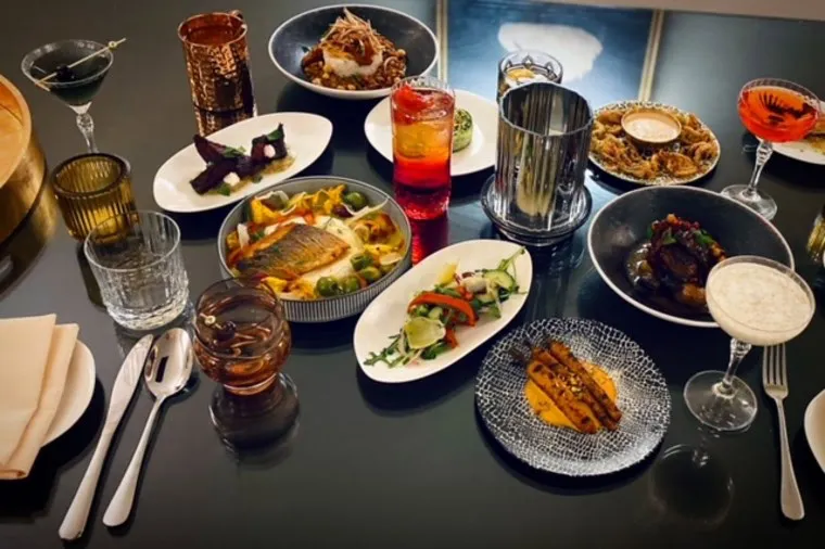 A table set with various gourmet dishes, including grilled fish, salad, pasta, roasted vegetables, and creative appetizers, accompanied by several colorful drinks and elegant glassware.