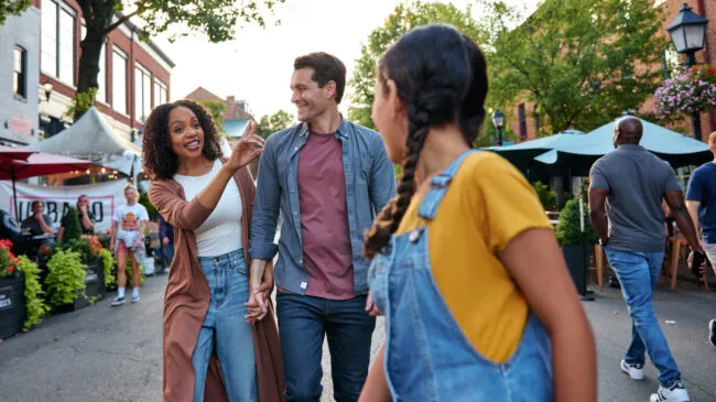 A smiling family walks together outdoors at a lively street market, with the woman gesturing as she talks, the man holding her hand, and a girl in front wearing overalls. Other people and market stalls are in the background.