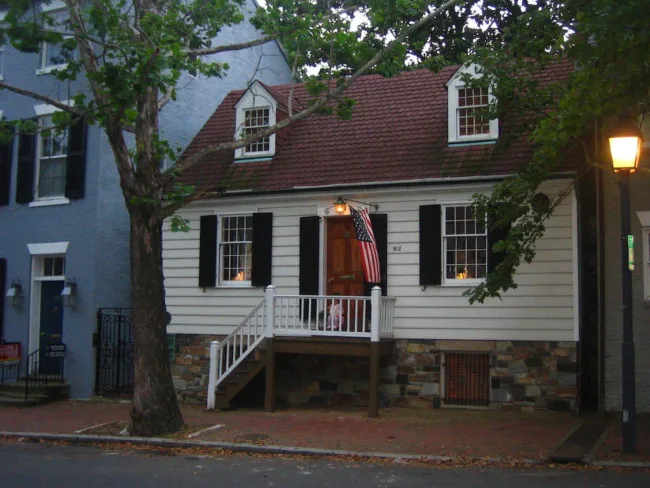 A small white house with a red roof, black shutters, and a stone foundation. An American flag hangs by the wooden front steps. A large tree and streetlamp are in front on a brick sidewalk.