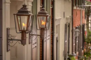 Two vintage gas lanterns with visible flames are mounted on the exterior wall of a building along a row of historic buildings. The image has a warm, nostalgic feel and shows architectural details and greenery in the background.