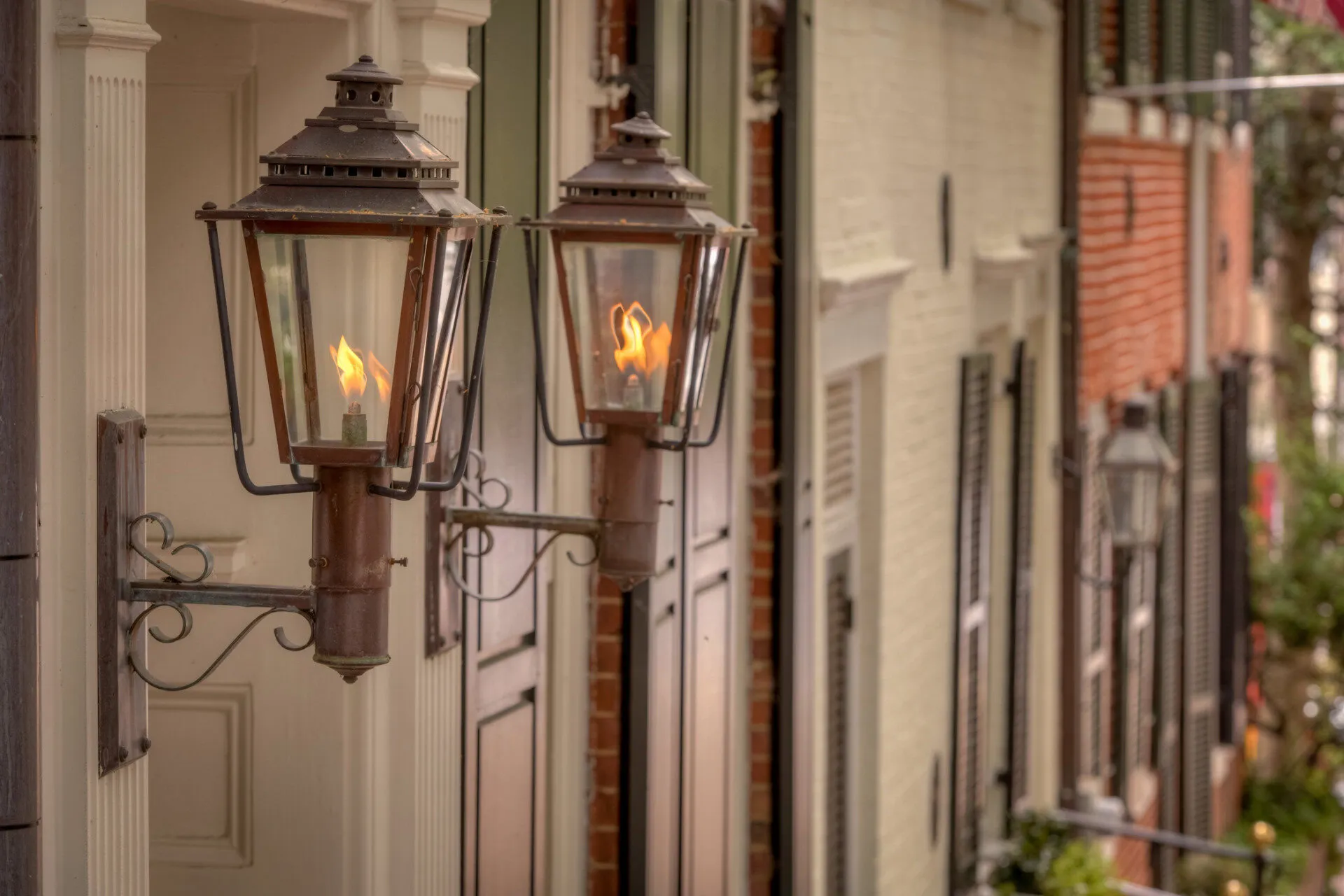 Two vintage gas lanterns with visible flames are mounted on the exterior wall of a building along a row of historic buildings. The image has a warm, nostalgic feel and shows architectural details and greenery in the background.