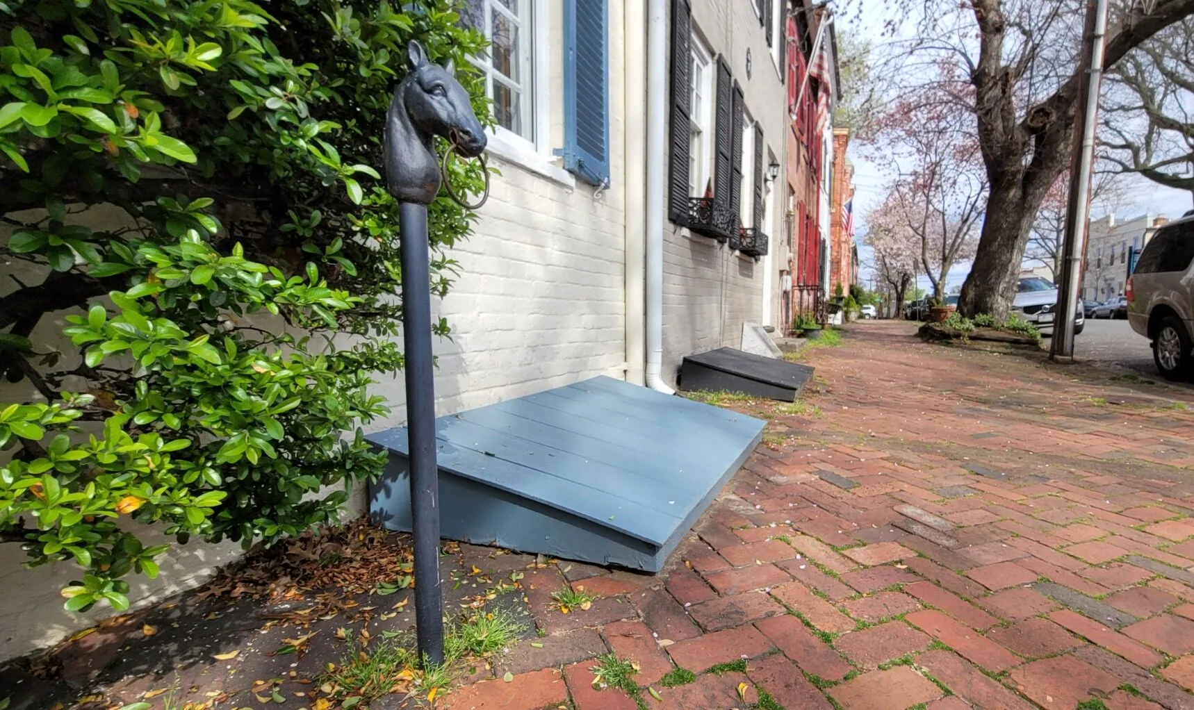 A black horse-head hitching post stands by a brick sidewalk and green bushes in front of a row of historic houses with outdoor cellar doors.