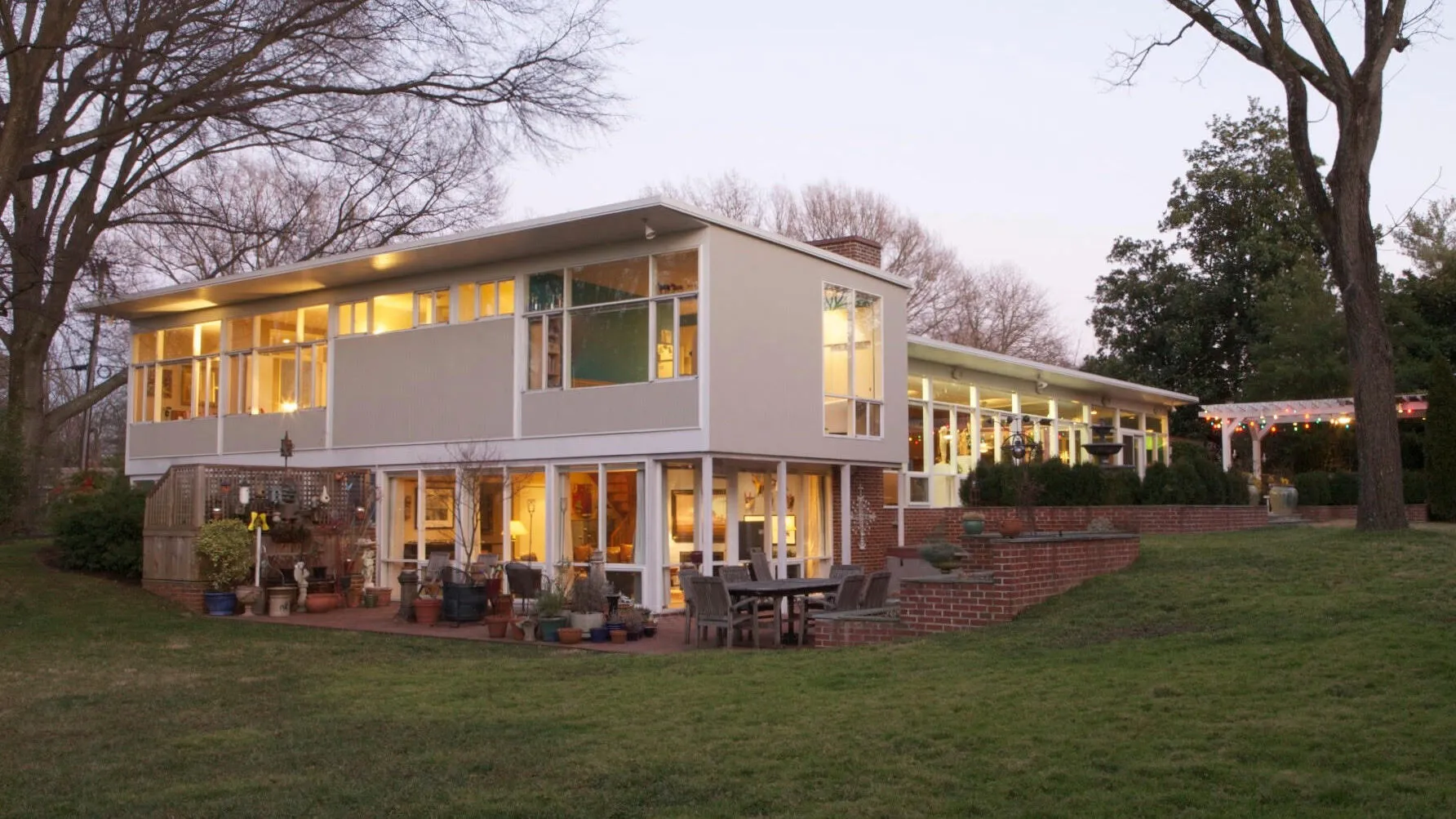 A modern, two-story house with large windows and glowing interior lights, surrounded by trees and a grassy yard. A brick patio with outdoor furniture and potted plants sits in the foreground.