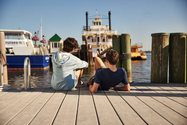 Two people sit on a wooden dock by the water, facing away. Boats and a ferry are docked ahead, and the sky is clear and blue. The scene appears relaxed and summery.