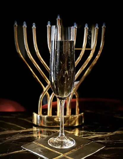 A glass of champagne sits on a dark, marble table with a gold menorah in the background, each candle unlit.