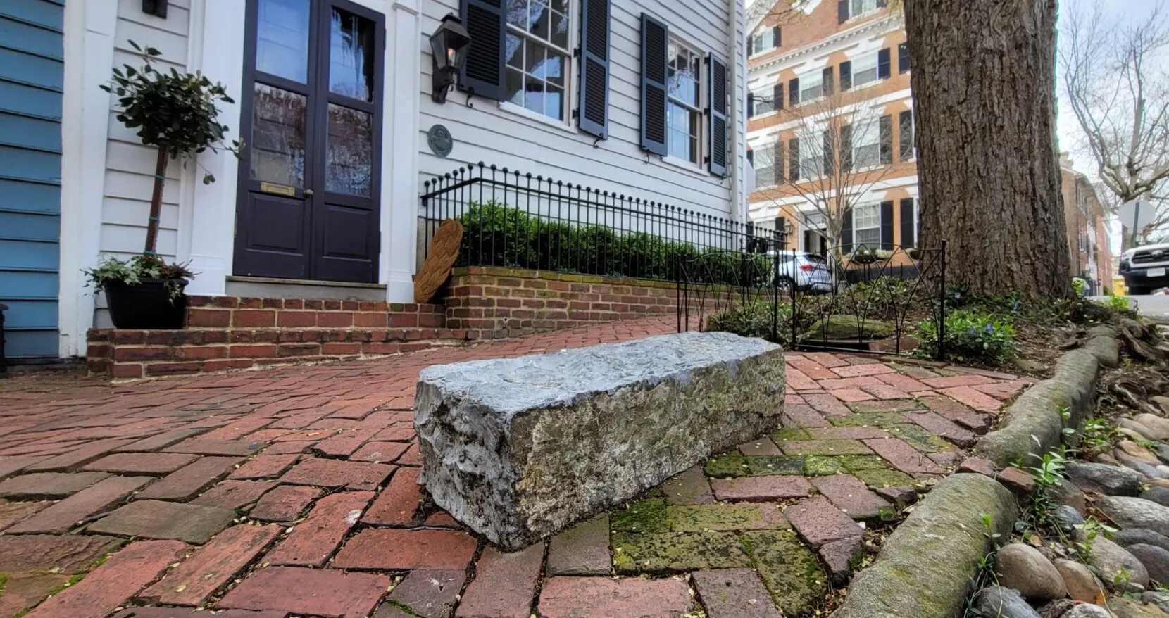 A rough stone block sits on a red brick sidewalk in front of a white house with black shutters and potted plants beside the entrance. A large tree and another historic building are visible in the background.
