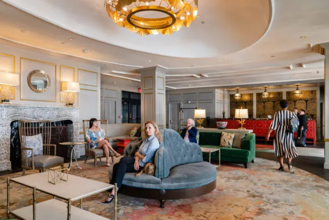 People sit and converse in a stylish hotel lobby with elegant furniture, gold accents, and a large chandelier. A woman sits on a curved sofa in the foreground, while others relax and talk around the room.