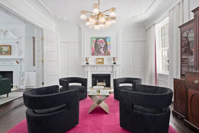 A modern living room with four curved black chairs around a white coffee table on a bright pink rug, a white fireplace, a colorful portrait above the mantel, and a gold chandelier overhead.