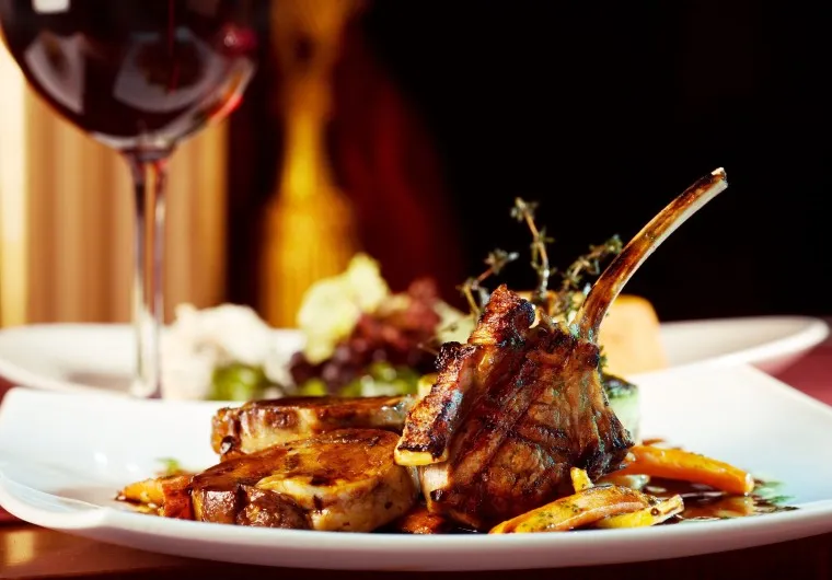 A close-up of grilled lamb chops served with roasted vegetables on a white plate, accompanied by a glass of red wine in the background. The meal is garnished with fresh herbs.