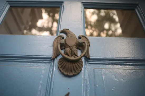 Close-up of a light blue door with a brass door knocker shaped like an eagle above a shell, and two glass window panes reflecting trees and sky in the background.