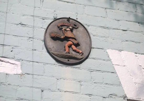A round metal plaque featuring a figure in a hat and boots carrying an object, mounted on a light blue painted brick wall with white accents.