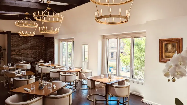 A bright, elegant restaurant dining area with round tables, cushioned chairs, large windows, modern chandeliers, a brick accent wall, and a framed painting. Tables are set with glasses, plates, and napkins.
