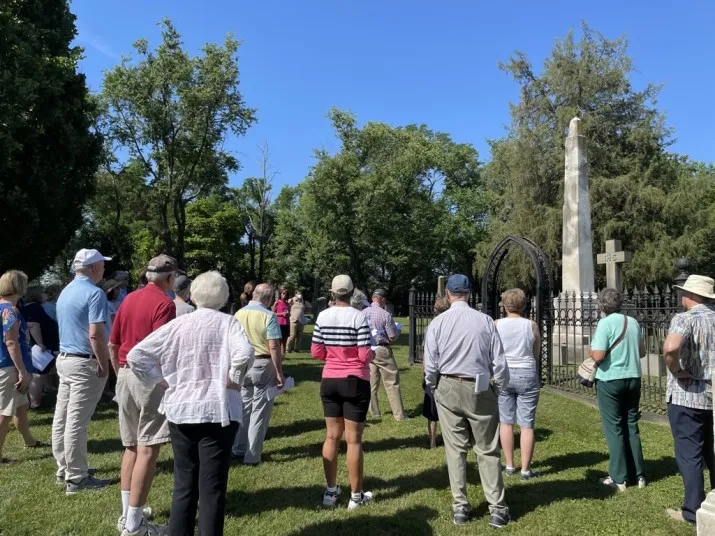 A group of people stand outdoors on a sunny day, listening to a guide near a fenced grave site with a tall obelisk monument and cross, surrounded by green grass and trees.