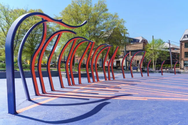 Sculptural bike rack with tall, curved blue and orange metal bars on a blue surface in an outdoor urban park, surrounded by trees and brick buildings under a clear sky.