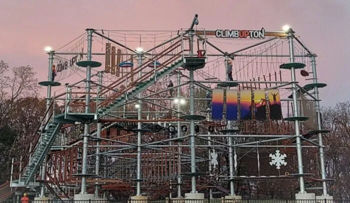 A large outdoor ropes and climbing adventure course called Climb Upton, with multiple platforms, ropes, bridges, and obstacles, set against a pink and purple evening sky. Trees are visible in the background.