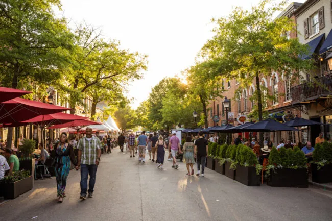 People stroll down a lively, sunlit street lined with outdoor cafes, red umbrellas, greenery, and brick buildings on both sides. Some sit and dine, while others walk and enjoy the pleasant atmosphere.