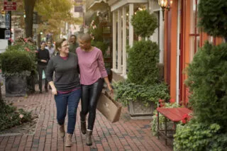 Two people walk arm-in-arm on a brick sidewalk, smiling at each other, holding a shopping bag. They pass by shops with greenery and flowers, while a small group walks behind them on an autumn day.