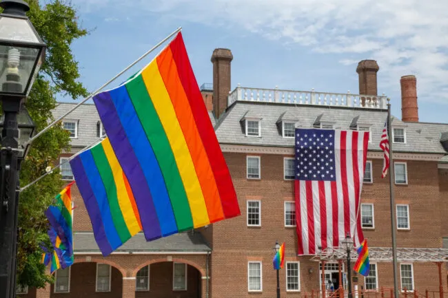 Large rainbow pride flags and American flags hang outside a brick building, with several small pride flags visible along the railing and a partly cloudy sky in the background.