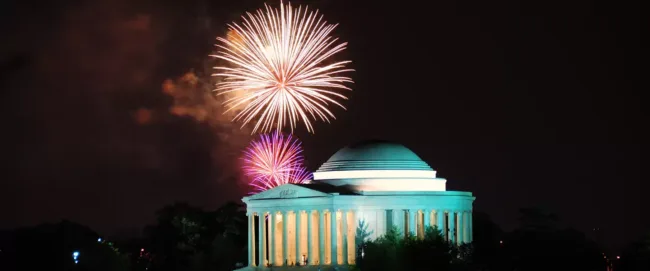 Fireworks light up the night sky above the illuminated Jefferson Memorial, featuring its domed roof and columns, surrounded by silhouettes of trees.
