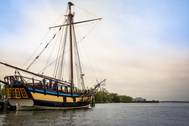 A historic sailing ship with tall masts and yellow trim is docked by the shore, with calm water and a distant treeline under a cloudy sky.