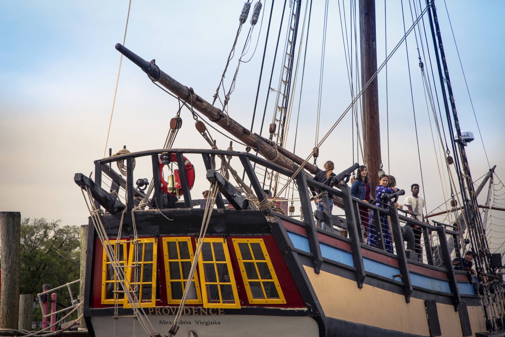 A group of people, including children, stand on the upper deck of a large historic sailing ship named Providence, docked at a pier. The ship features tall masts, rigging, and yellow-trimmed windows.