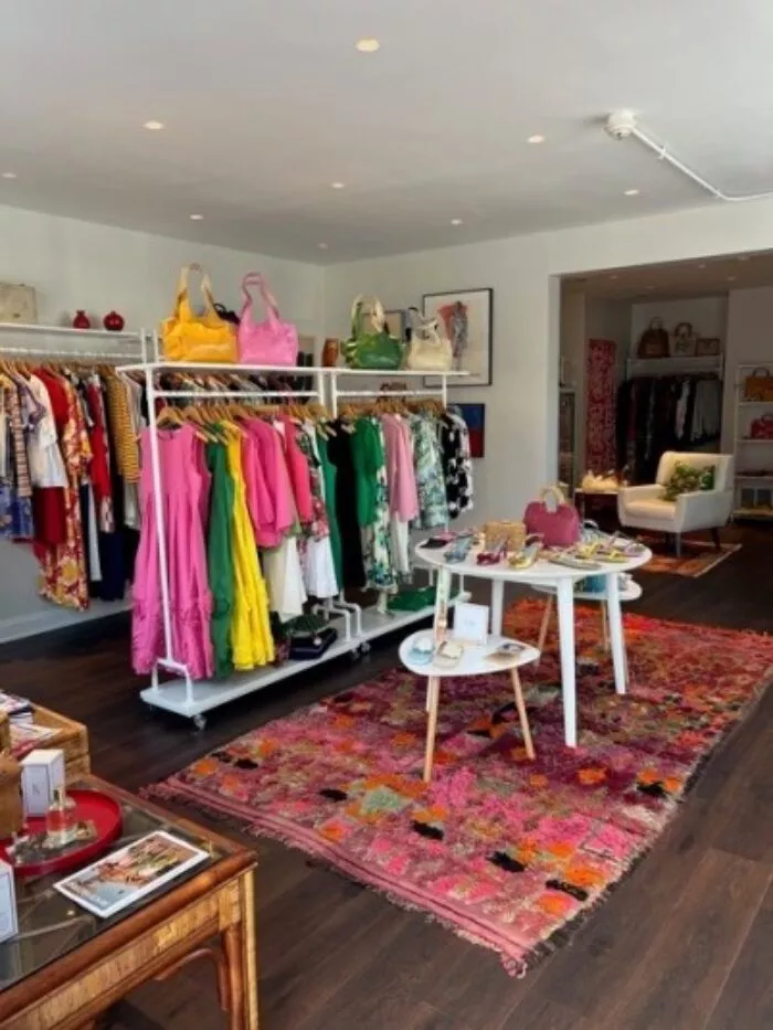 A boutique with colorful dresses on racks, bright handbags on shelves, and a table displaying accessories. A patterned rug covers the dark wood floor, and a white couch sits in the back corner.