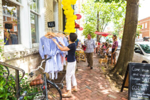 A woman browses light blue dresses on a rack outside a boutique with a summer sale sign. Yellow balloons, a chalkboard, and people with dogs are on a lively, tree-lined brick sidewalk.