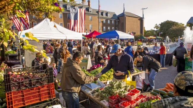 A busy outdoor farmers market with people shopping for fresh produce at various vendor stalls. Colorful tents and American flags are visible, with a fountain and brick buildings in the background.