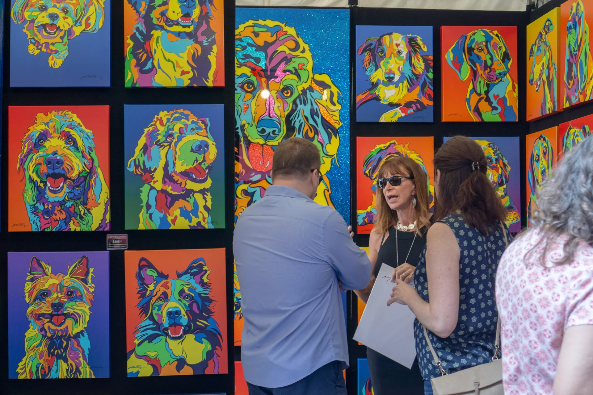 Three people stand and talk in front of a display of colorful, pop art-style dog portraits at an outdoor art show. The paintings feature various dog breeds in bright, vibrant colors.