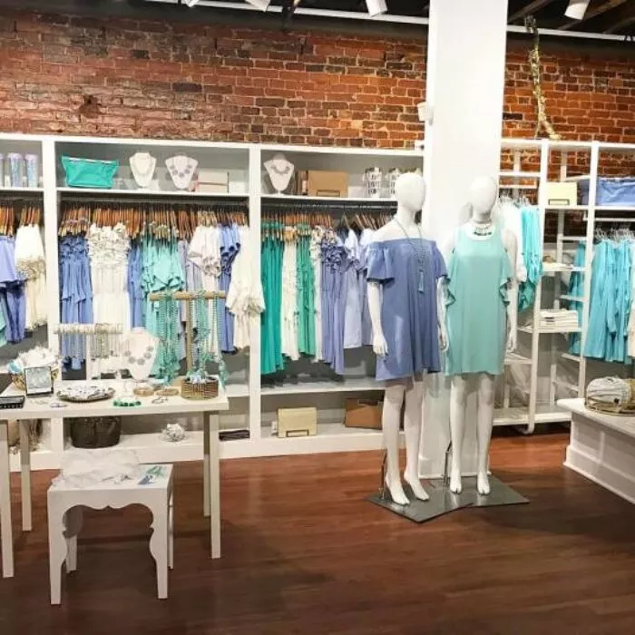 A boutique clothing store with brick walls, wooden floors, and racks of pastel blue and teal dresses. Two mannequins display blue dresses, with jewelry and accessories arranged on nearby tables.