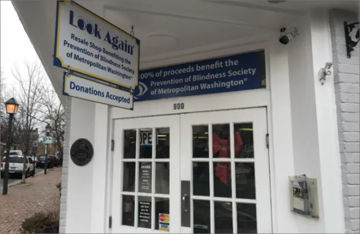 A white storefront with signs reading Look Again and 100% of proceeds benefit the Prevention of Blindness Society of Metropolitan Washington. A donation box and a door with store hours are visible.