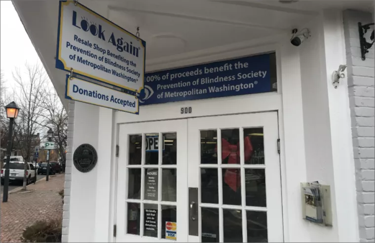 A white storefront with signs reading Look Again and 100% of proceeds benefit the Prevention of Blindness Society of Metropolitan Washington. A donation box and a door with store hours are visible.