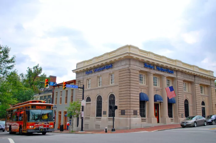 A red trolley bus drives past a historic brick bank building with blue awnings and an American flag on a corner in a small town, with street signs and trees visible.