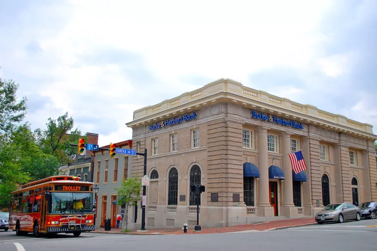 A red trolley bus drives past a historic brick bank building with blue awnings and an American flag on a corner in a small town, with street signs and trees visible.