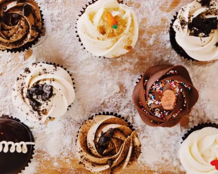 Assorted cupcakes with swirled frosting, chocolate, cookie crumbles, and colorful sprinkles are arranged on a floured wooden surface.
