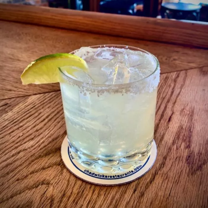 A margarita served in a salt-rimmed glass with ice and a lime wedge, sitting on a coaster atop a wooden table.