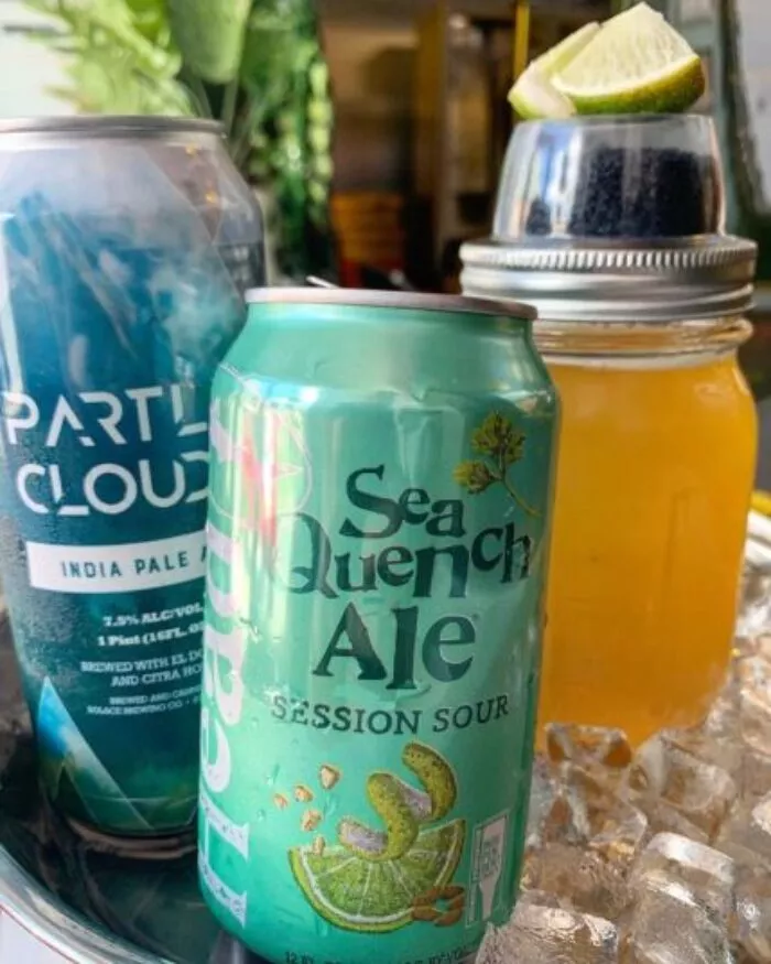 Two cans of craft beer, one labeled Sea Quench Ale and the other Party Cloudy, sit on ice next to a mason jar filled with a yellow drink, garnished with a lime wedge. Green plants are in the background.