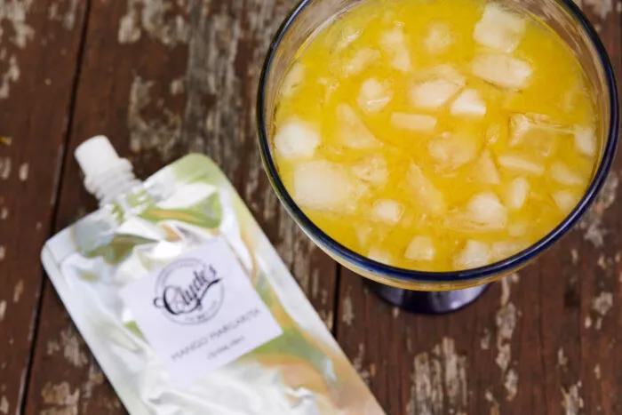 A cocktail glass filled with an orange-yellow drink and ice sits on a wooden surface next to a silver pouch labeled “Clyde’s Mango Pineapple.”.