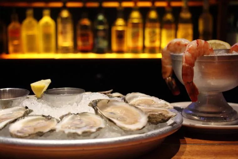 A platter of fresh oysters on ice with lemon and sauce, next to a bowl filled with shrimp cocktail and lemon wedges; bottles of alcohol are blurred in the background.