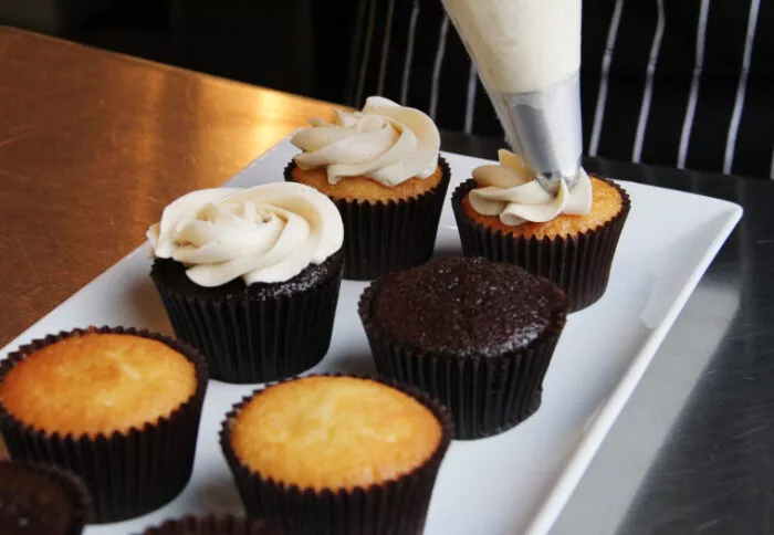 A person pipes vanilla frosting onto cupcakes, both chocolate and vanilla, arranged on a white rectangular plate. The person wears a black apron with white stripes.