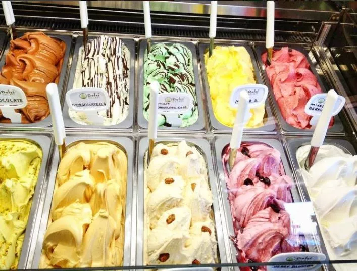 A display of colorful gelato flavors in a shop, each in a metal container with a spatula. Flavors include chocolate, stracciatella, pistachio, lemon, strawberry, and more, each with a small label.