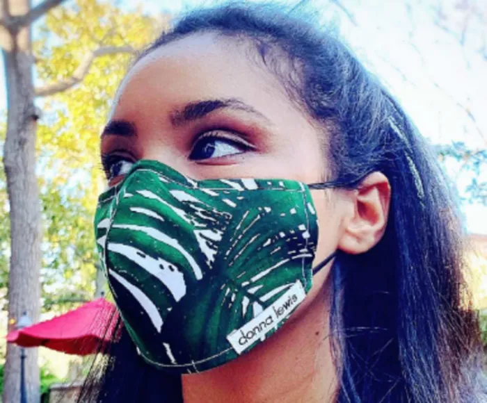 A woman with long dark hair wears a green face mask with a white leaf pattern and a “donna lewis” label, standing outdoors among trees and sunlight.