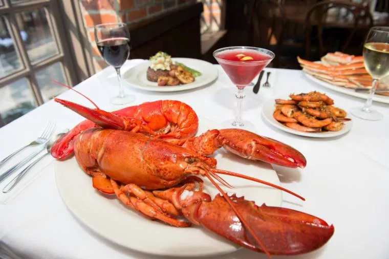 A large cooked lobster on a white plate is set on a table with wine, a cocktail, shrimp, crab legs, and a steak dish in the background, all arranged on a white tablecloth.