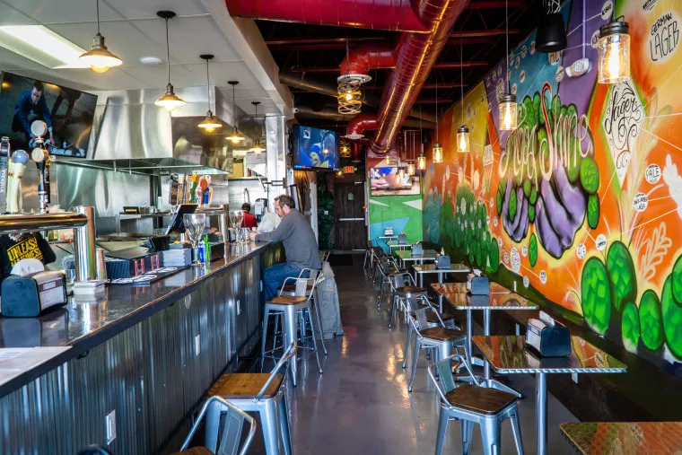 A colorful restaurant interior with vibrant wall graffiti, a long bar on the left, metal stools, and small tables. One person sits at the bar while several TVs and hanging lights decorate the space.