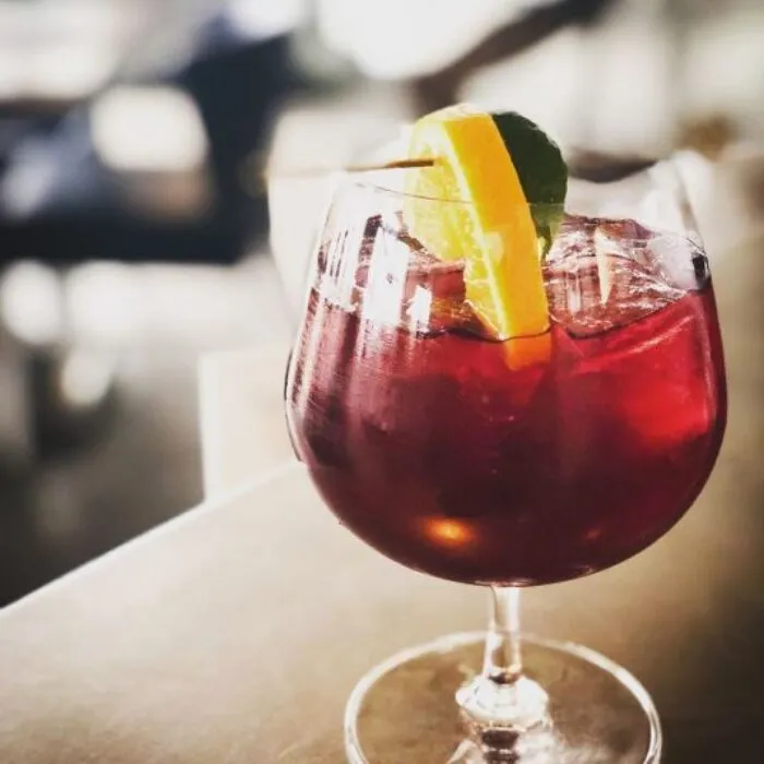 A large glass filled with a deep red cocktail, ice cubes, and garnished with a slice of lemon and lime on a skewer, sits on a table with a blurred background.