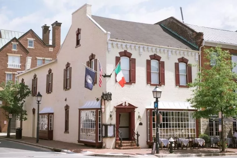 A white brick building with burgundy trim stands on a street corner, displaying three flags and featuring large windows, outdoor seating with white tablecloths, and a tree to the right.