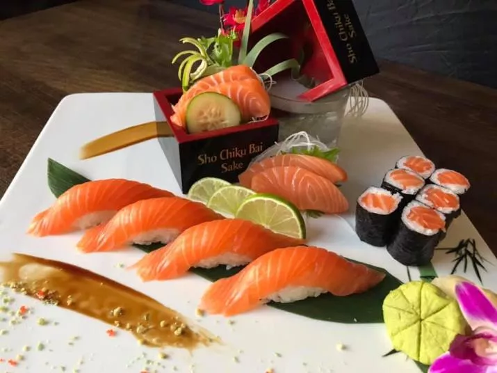 A platter of assorted salmon sushi, including nigiri and maki rolls, garnished with lime slices, a cucumber slice, and a decorative flower, served on a white plate with sauce and colorful edible accents.