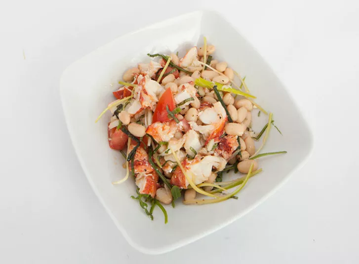 A square white bowl filled with a salad of white beans, tomato pieces, lobster meat, and assorted microgreens on a plain white background.