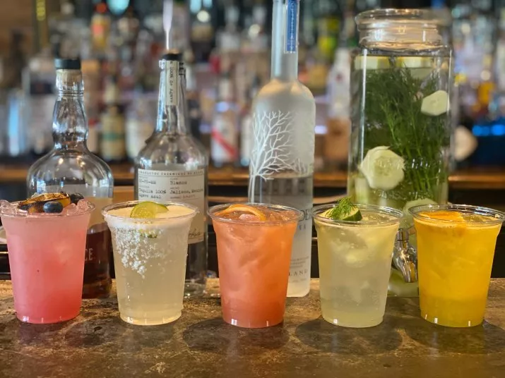 Six colorful cocktails in plastic cups are lined up on a bar counter, each garnished with fruit or herbs. Behind them are bottles of liquor and a large jar with cucumber and herbs infused in water.