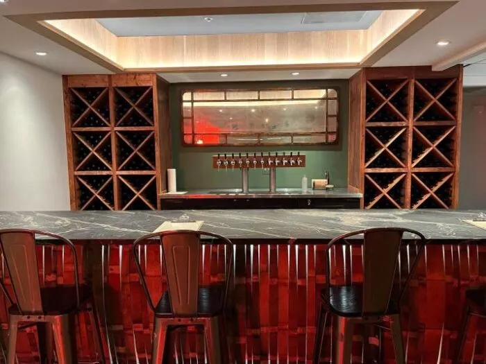 A modern bar with a dark marble countertop, metal bar stools, wooden wine racks filled with bottles, and a set of beer taps beneath a vintage-style mirror. Warm lighting creates a cozy atmosphere.